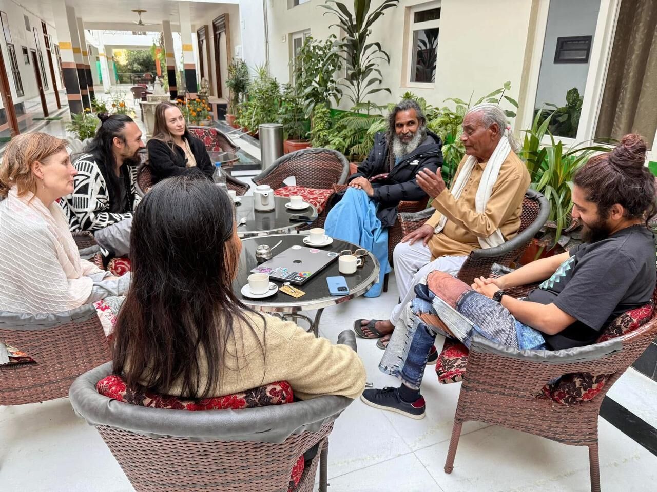 A group of people seated around a round table in an outdoor setting with wicker furniture and potted plants.