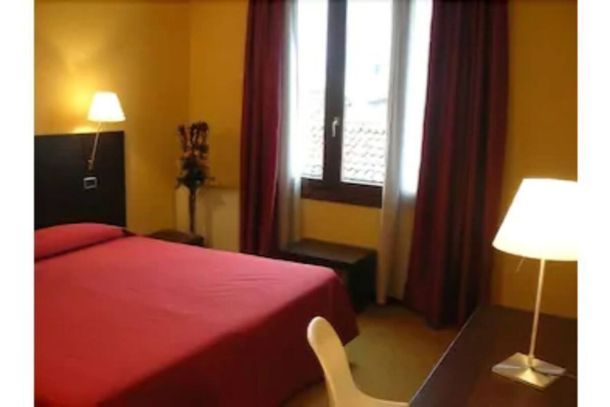 Une chambre d’hôtel avec un lit, un bureau, une chaise, une lampe et une fenêtre avec des rideaux.