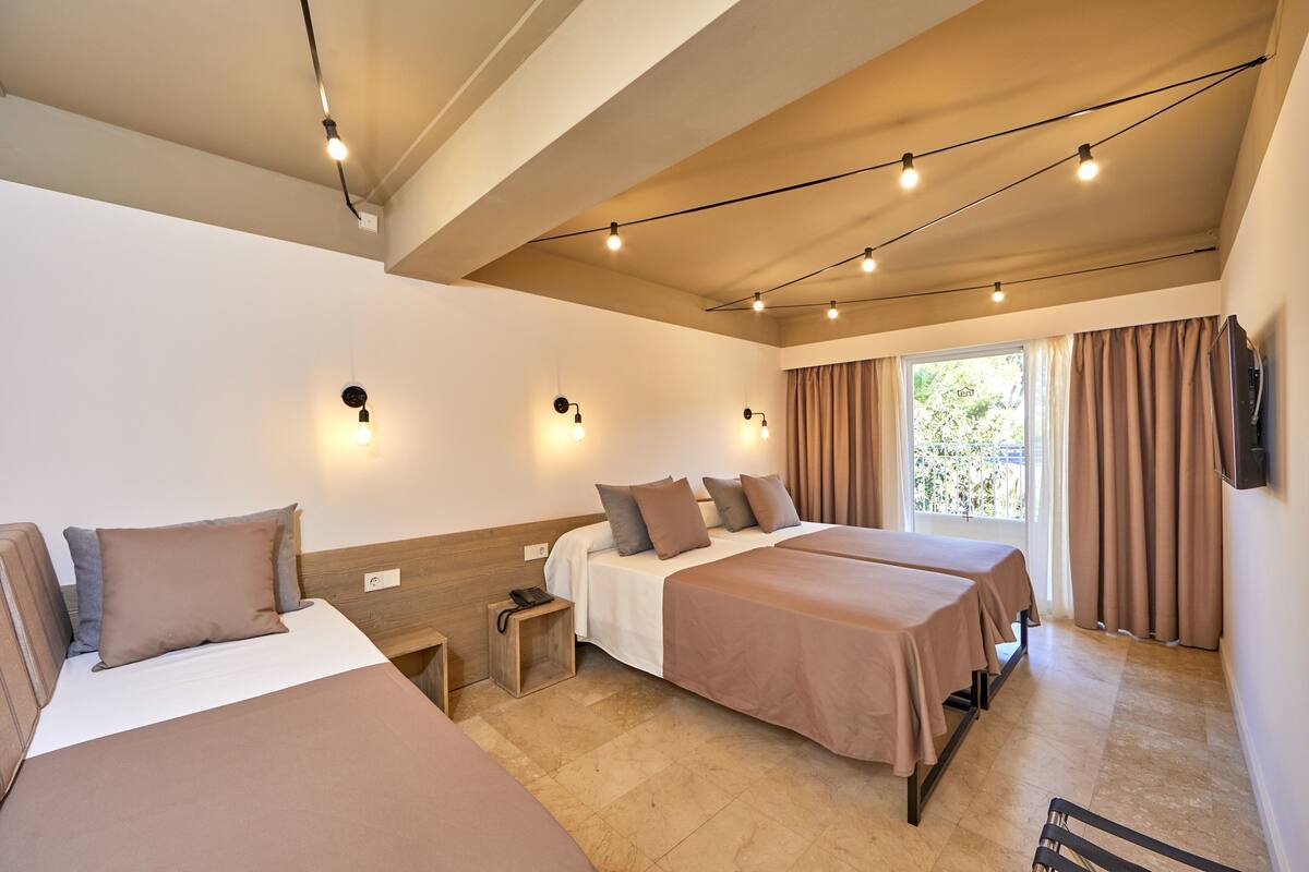 A hotel room with two beds, a small bedside table, a wall-mounted TV, and a window with curtains.