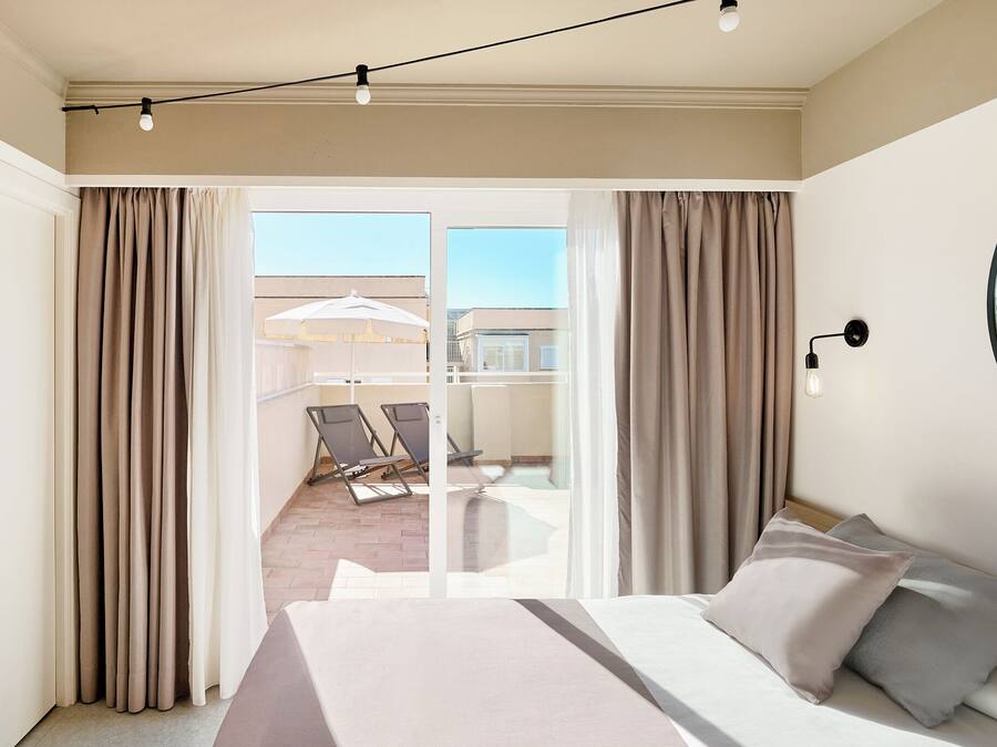 A hotel room with a bed, curtains, a balcony with chairs, and a view of buildings.