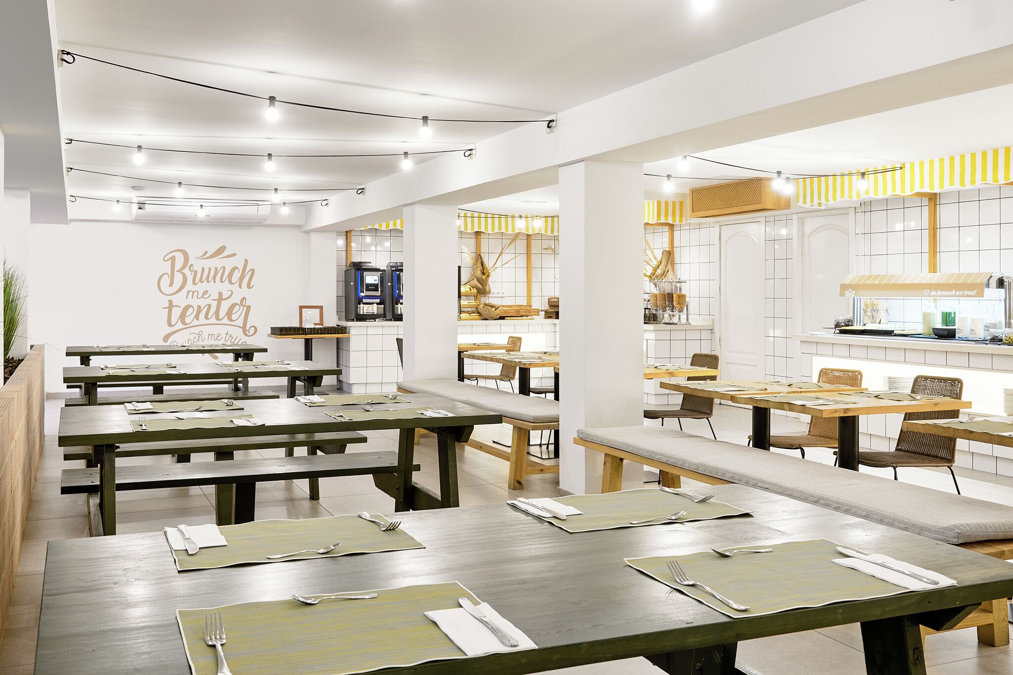Un restaurante moderno con mesas y bancos de madera, una zona de cocina y una pared con el letrero 'Brunch me tender'.