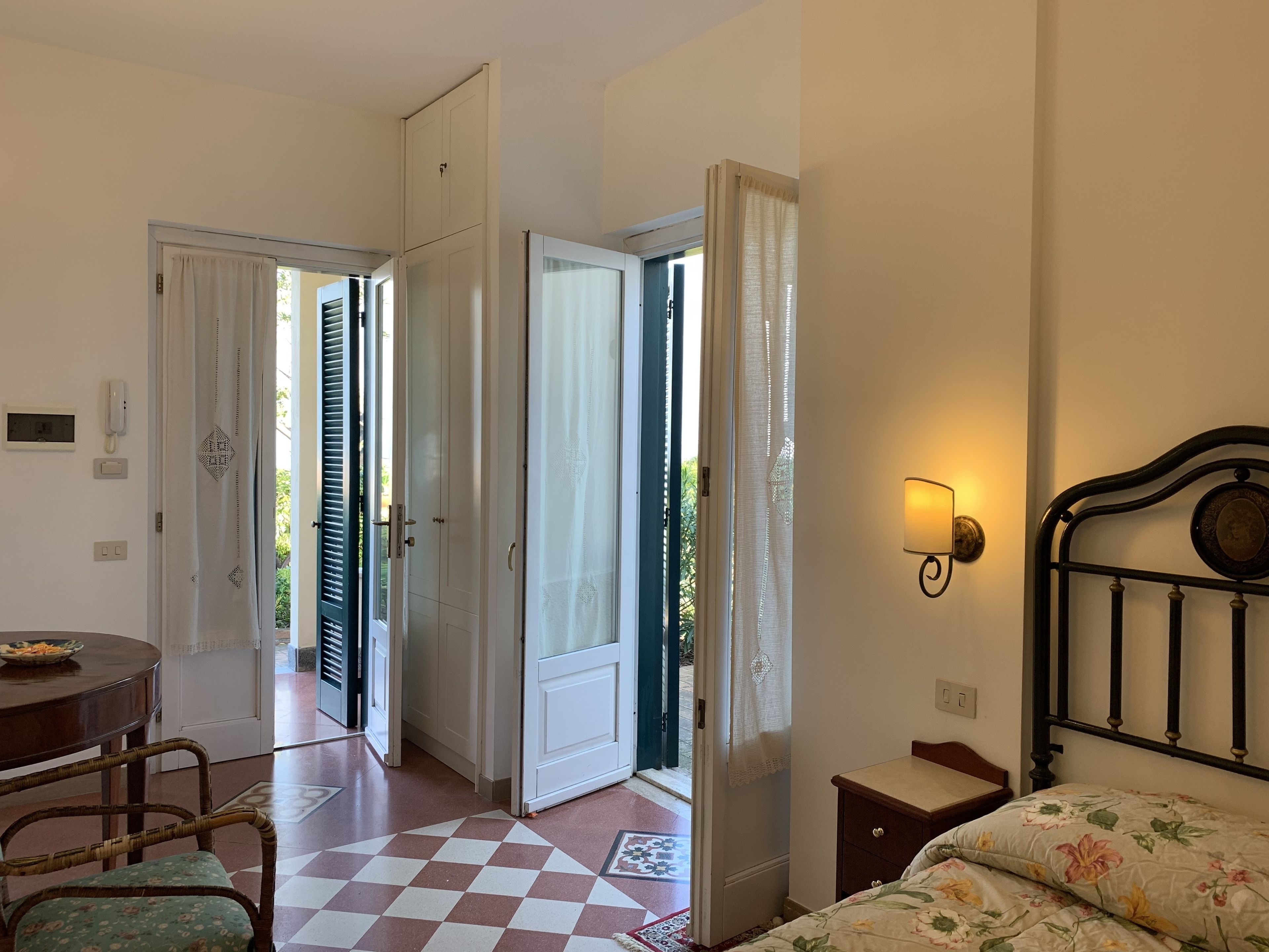 Une chambre d’hôtel avec un sol à carreaux, un lit recouvert d’une literie à motifs floraux, une petite table de chevet, une lampe fixée au mur et une vue sur l’extérieur par une porte ouverte.
