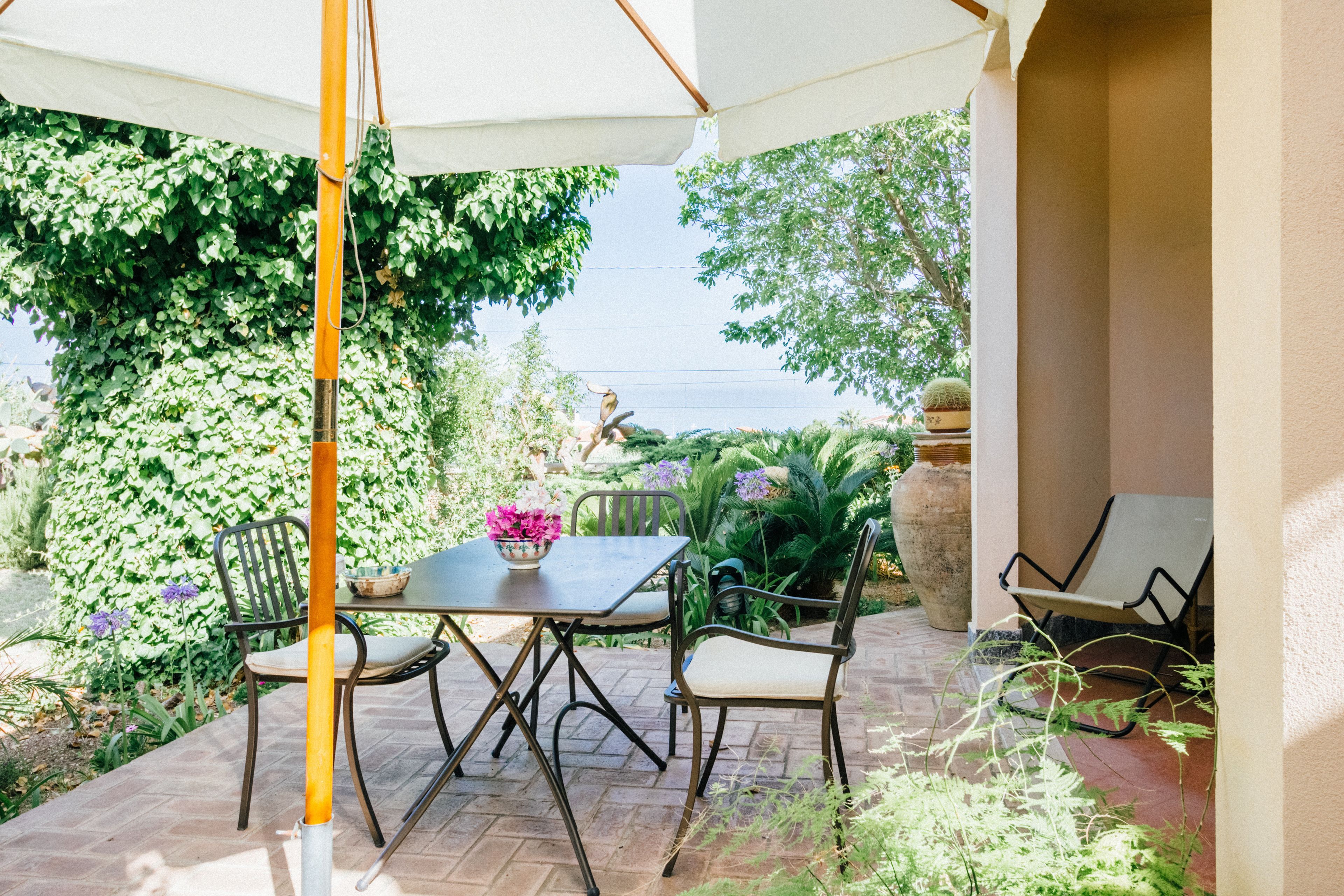 Eine Terrasse im Freien mit Tisch und Stühlen, umgeben von Grünpflanzen und einem großen Sonnenschirm.