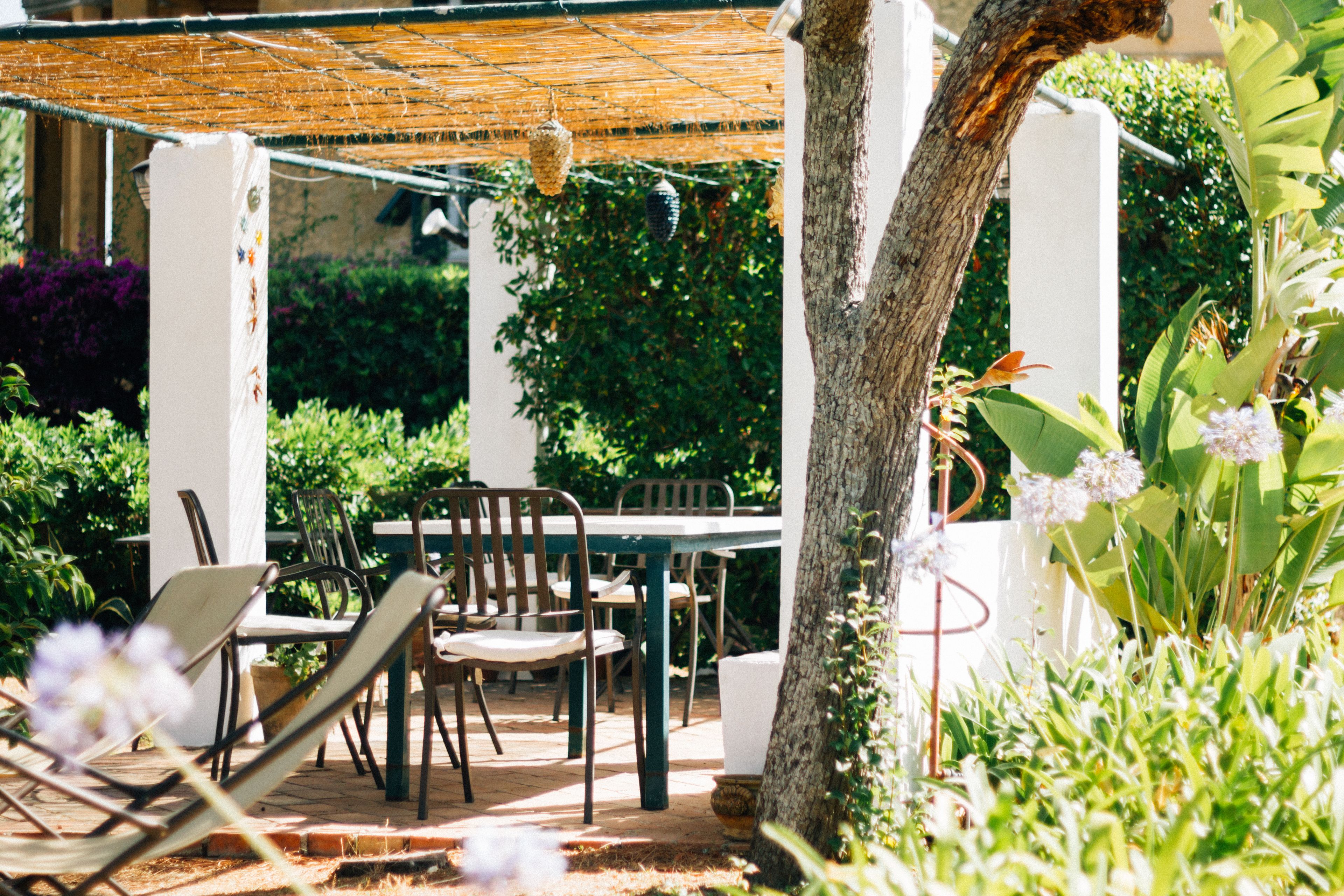 Um pátio externo com mesa e cadeiras, cercado por vegetação e uma pérgula.
