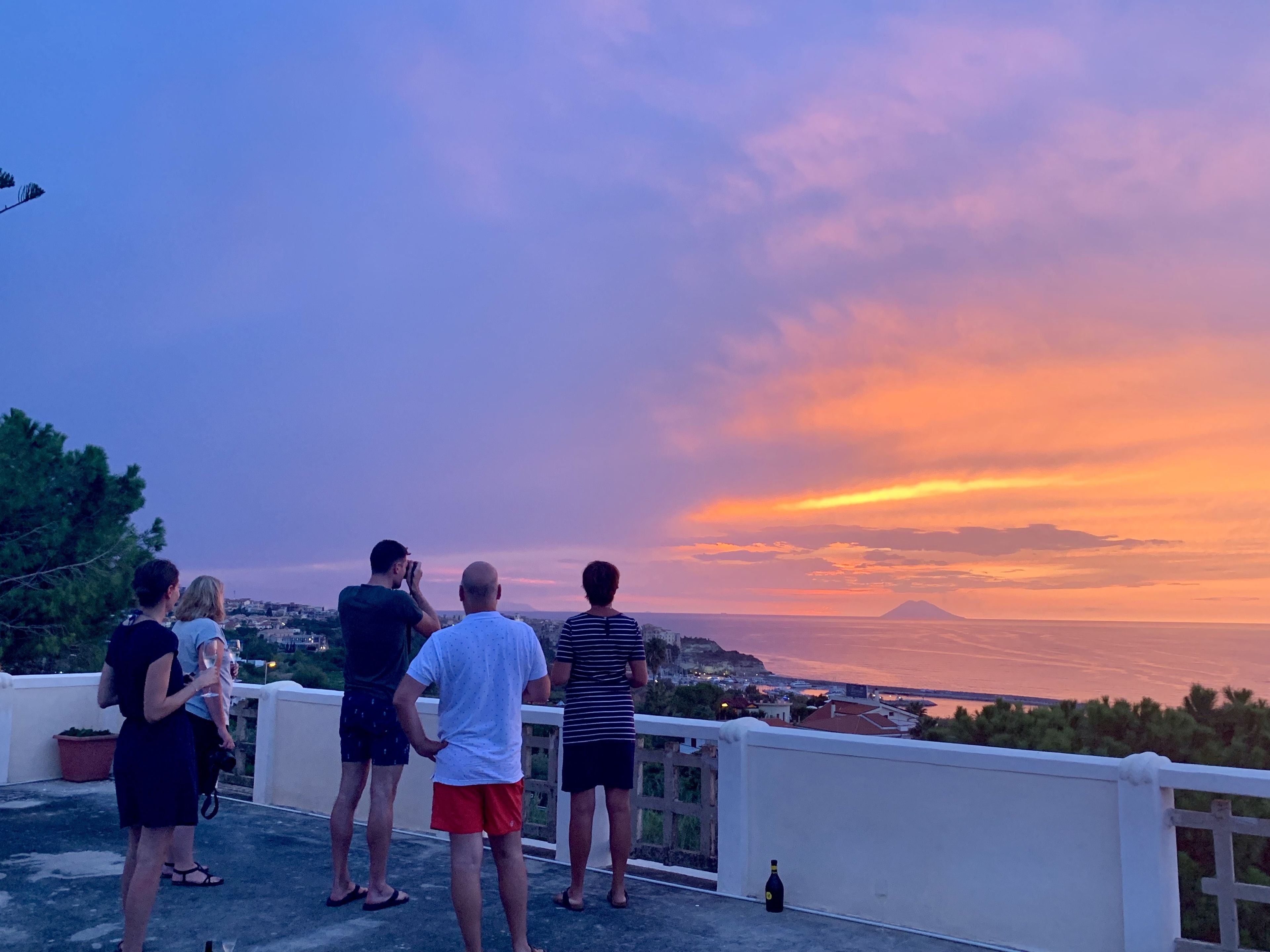 Eine Gruppe von Personen steht auf einem Balkon und blickt auf eine malerische Landschaft bei Sonnenuntergang.