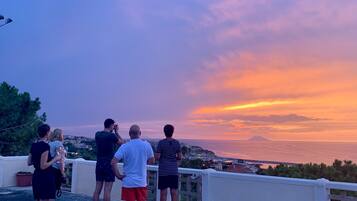Eine Gruppe von Personen steht auf einem Balkon und blickt auf eine malerische Landschaft bei Sonnenuntergang.