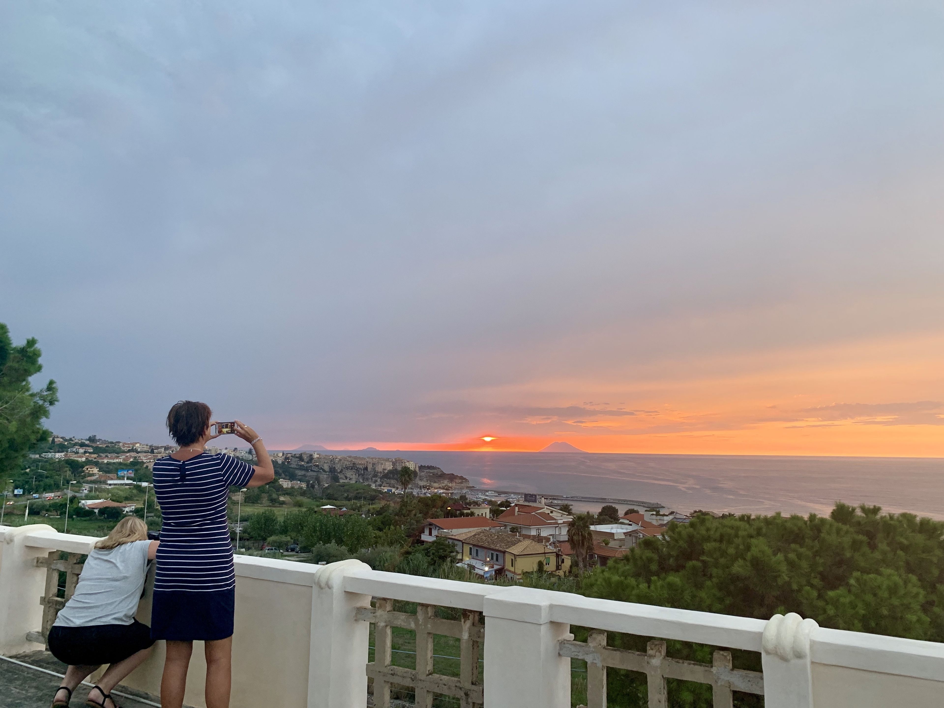 A balcony overlooking a coastal town with a sunset view, a mountain in the distance, and a person taking a photo.