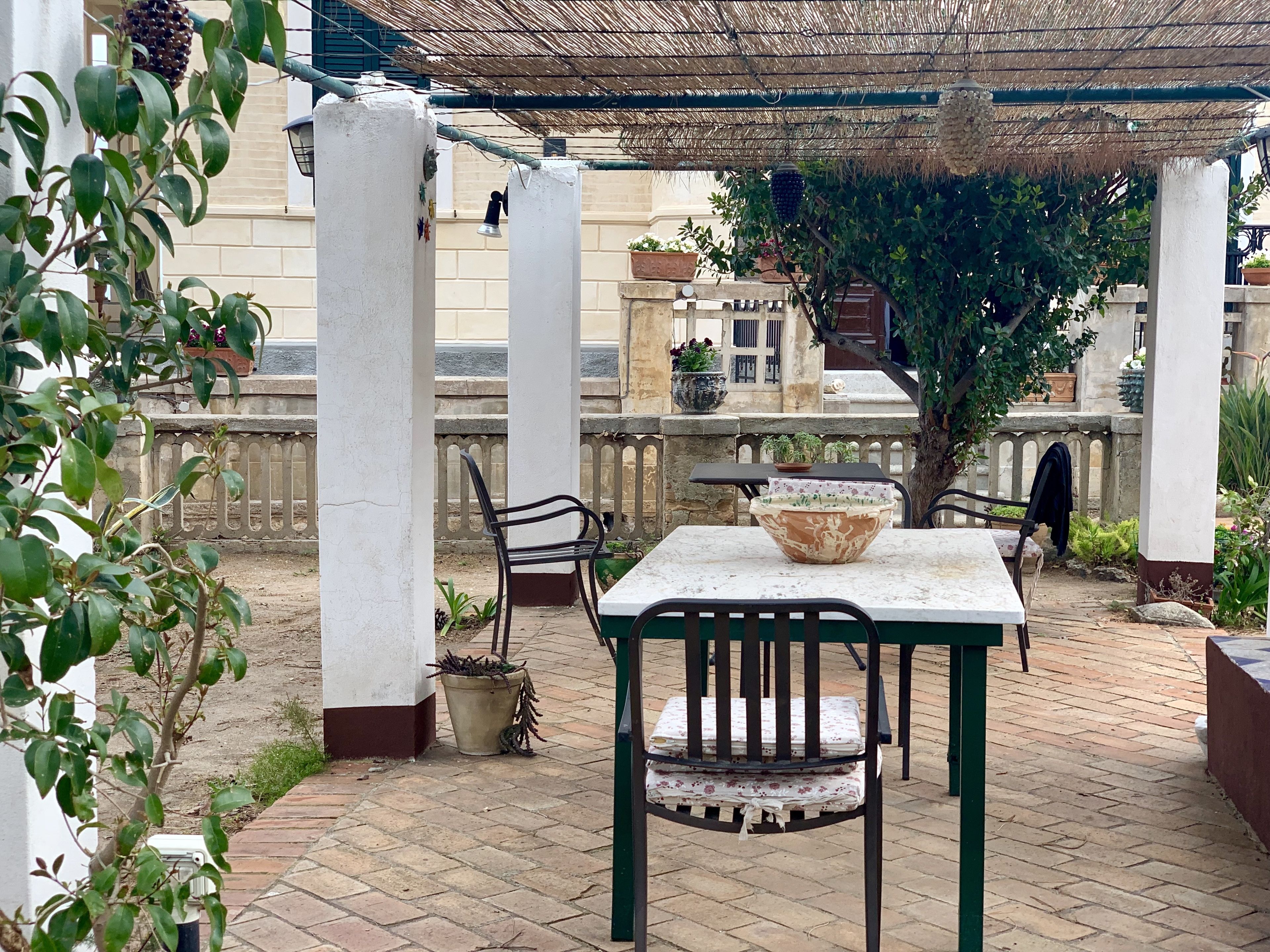 En udendørs terrasse med et bord og stole, omgivet af krukker med planter og en tagkonstruktion dækket med strå.
