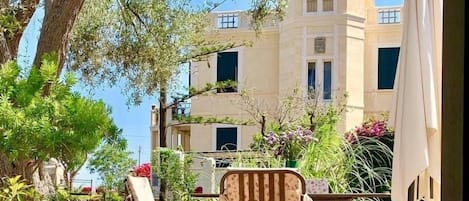 Un patio al aire libre con dos sillas y una mesa, rodeado de follaje y un edificio al fondo.