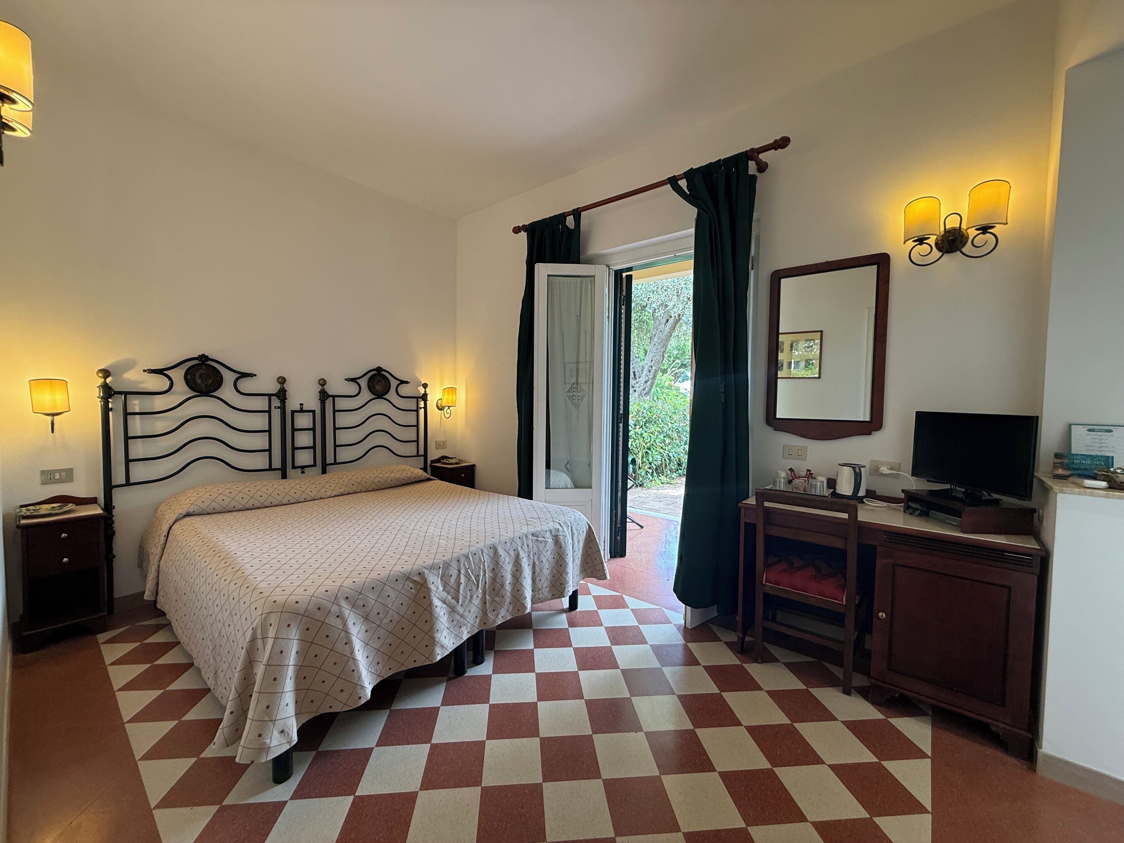 A bedroom with a checkered floor, a bed with a patterned bedspread, a nightstand, a dresser, a television, and a window with curtains.