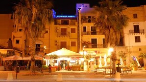 Escena urbana nocturna con un café al aire libre en el centro, palmeras y edificios con letreros iluminados.
