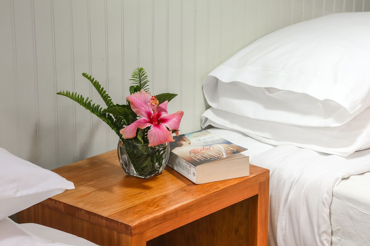 A wooden bedside table with a book titled "Rachel's Holiday" and a vase with pink flowers.
