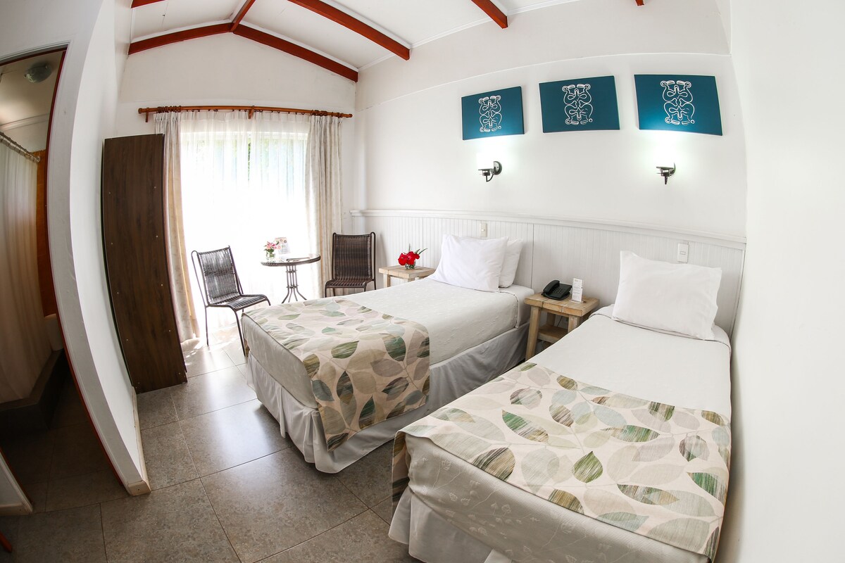 A hotel room with two beds, a small table with a chair, a window with curtains, and wall art.