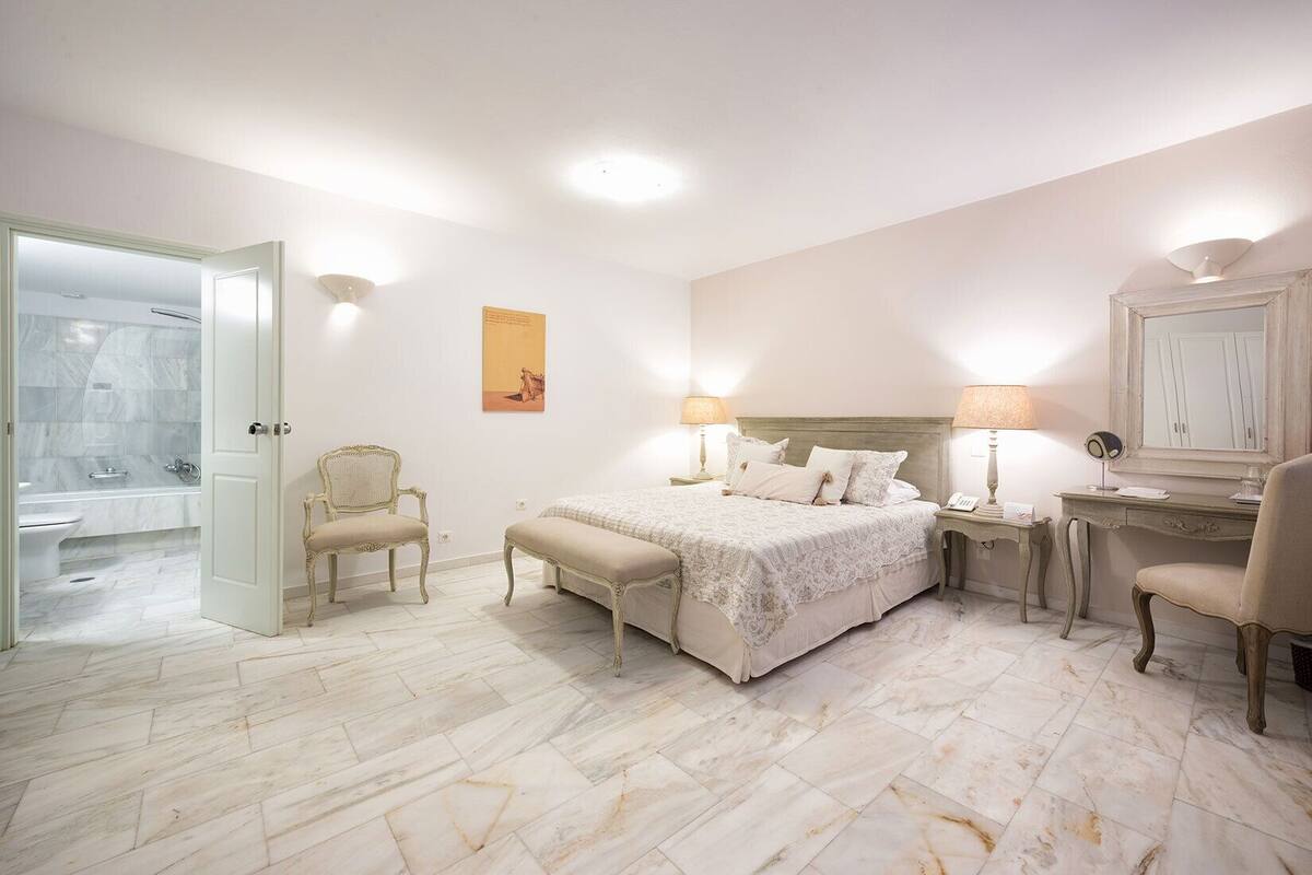 Un dormitorio con cama, mesitas de noche, una silla, un escritorio y un baño visible a través de una puerta abierta.