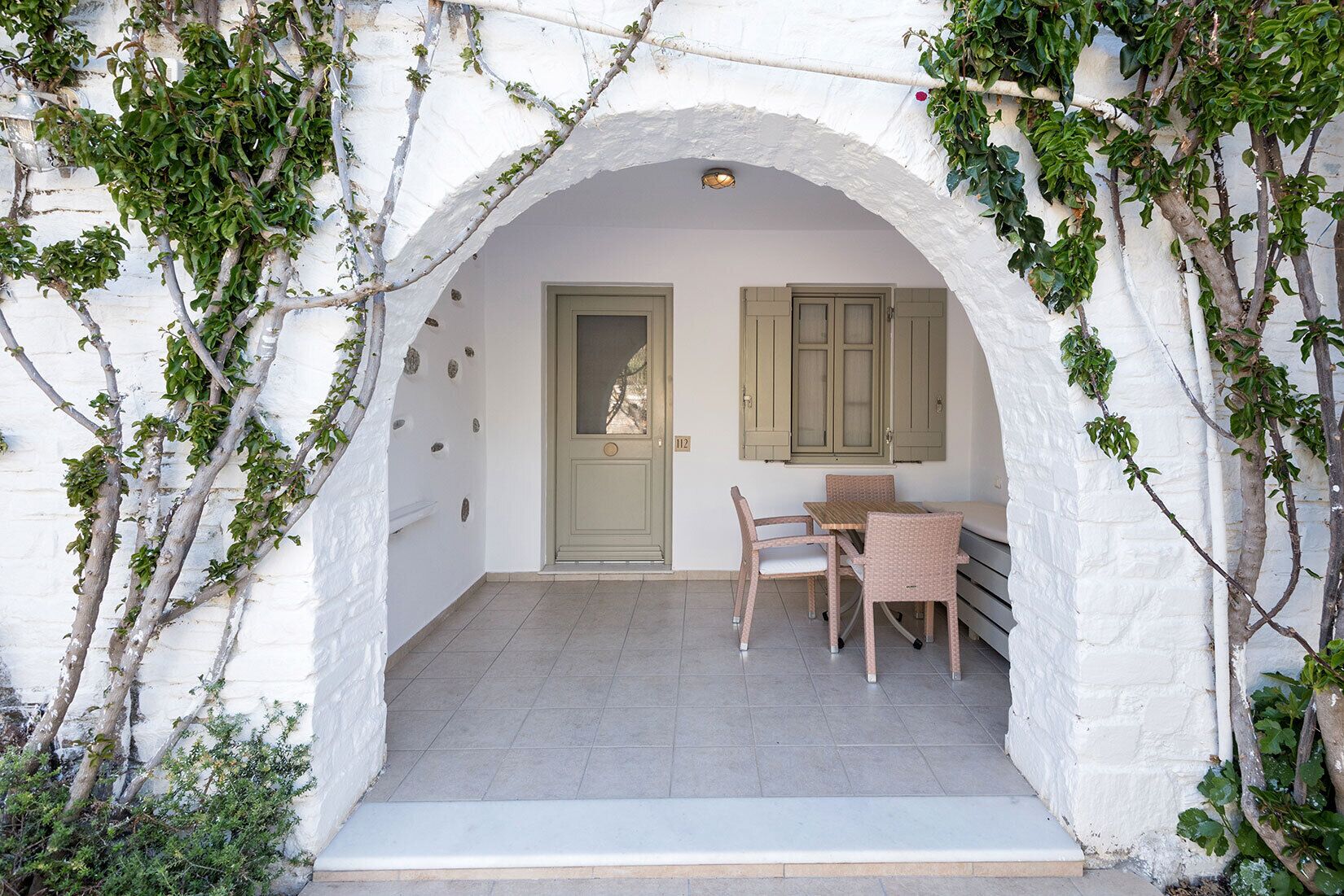 A covered outdoor seating area with a table and chairs, a door, and a window with shutters.