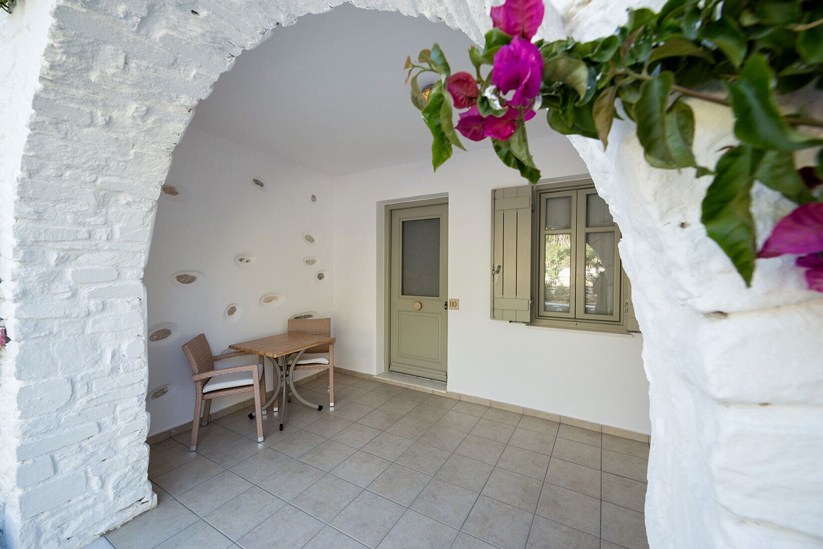 Una habitación con pared de ladrillo blanco, una mesa de madera con dos sillas, una puerta y un ventanal con contraventanas.
