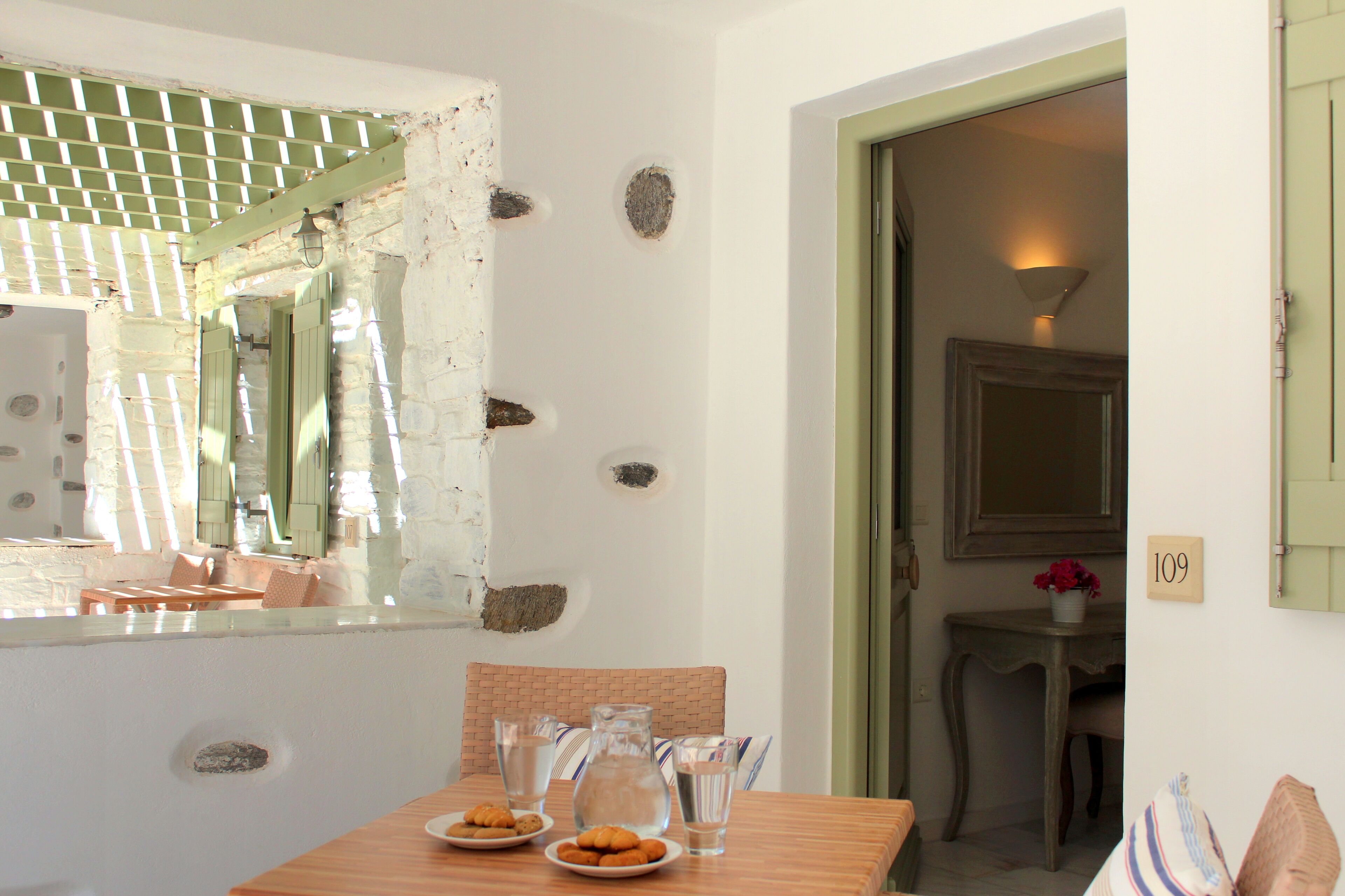 A dining area with a table set for two, a pitcher, glasses, and plates of food. There is a window with a view of the outside, a door leading to another room, and a number sign on the wall.