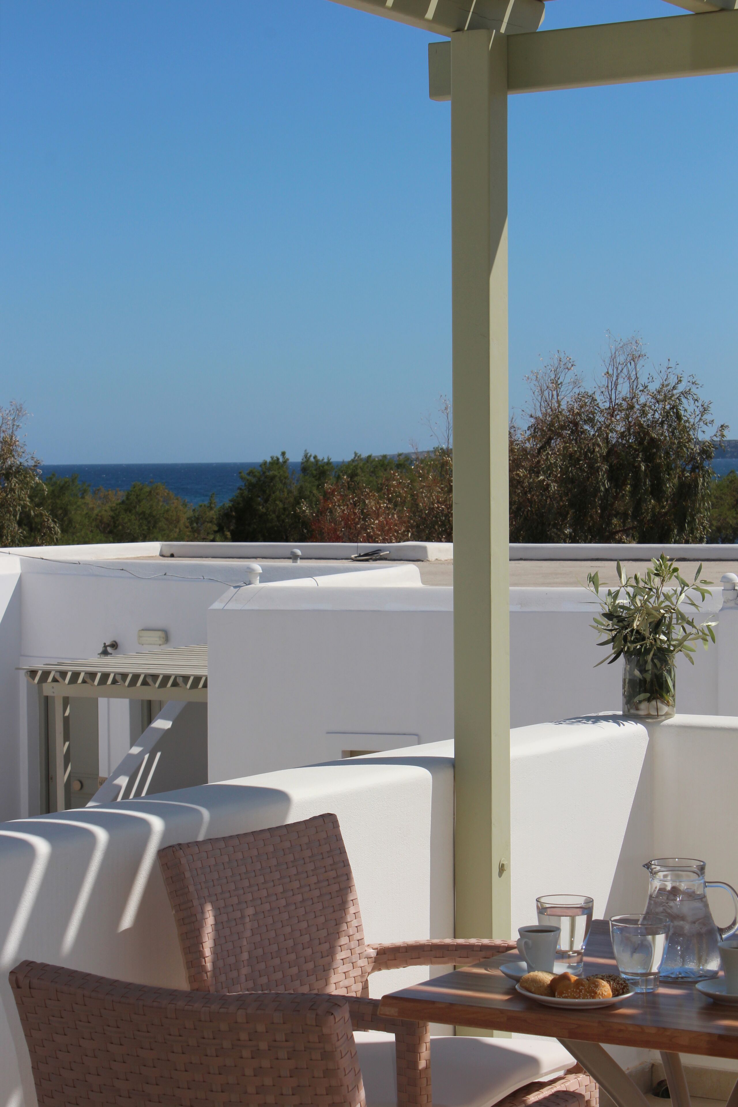 A terrace with white furniture, a table with a pitcher, cups, and a plate of cookies, and a view of the sea.