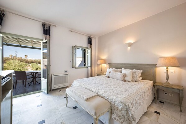 A bedroom with a large bed, a desk with a chair, a window with curtains, and a view of the outdoors. - Saint Andrea Sea Side Resort - Adults Only (Paros)