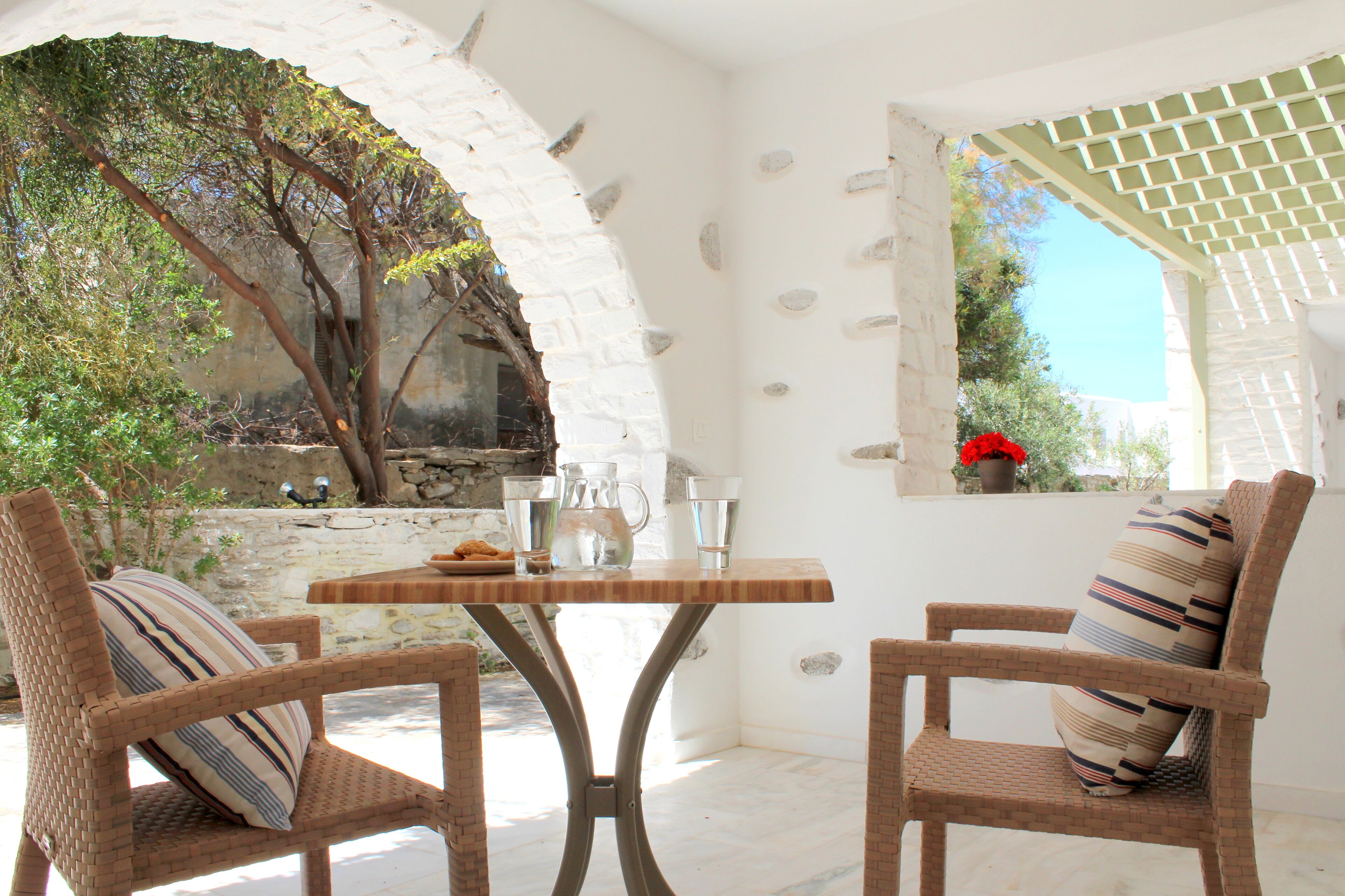 Una habitación con sillas de mimbre, una mesa de madera y vistas a árboles y un muro de piedra.