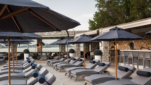 A sunlit outdoor lounge area with multiple lounge chairs and umbrellas, stone walls, and a view of the water. - Saint Andrea Sea Side Resort - Adults Only (Paros)