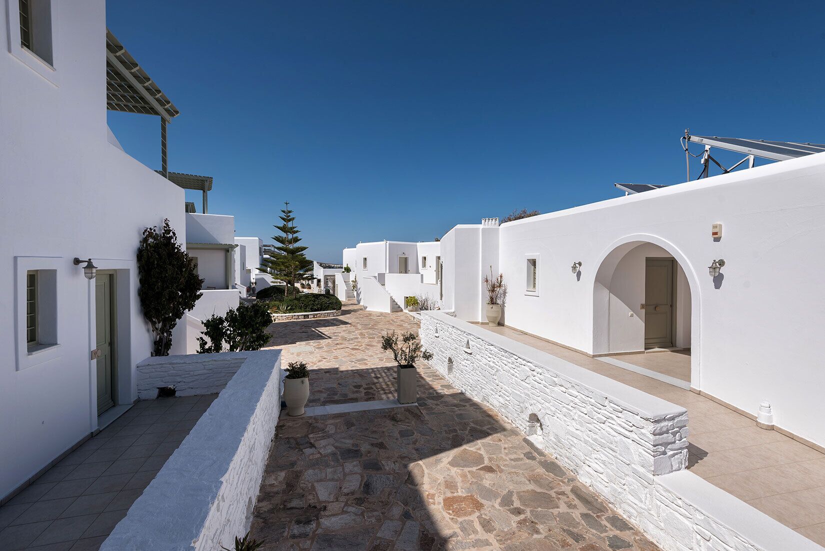 A narrow, stone-paved courtyard with white buildings, arched doorways, and a clear blue sky.