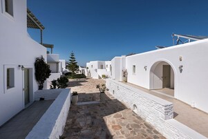 A narrow, stone-paved courtyard with white buildings, arched doorways, and a clear blue sky.