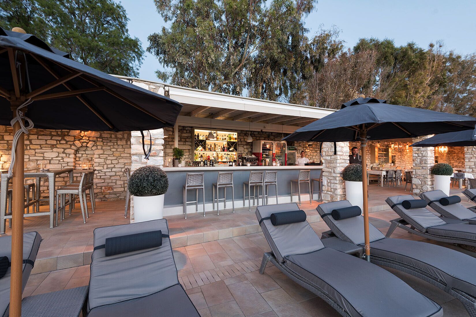 A bar area with a stone wall, bar stools, and a counter. There are lounge chairs with umbrellas and a potted plant.