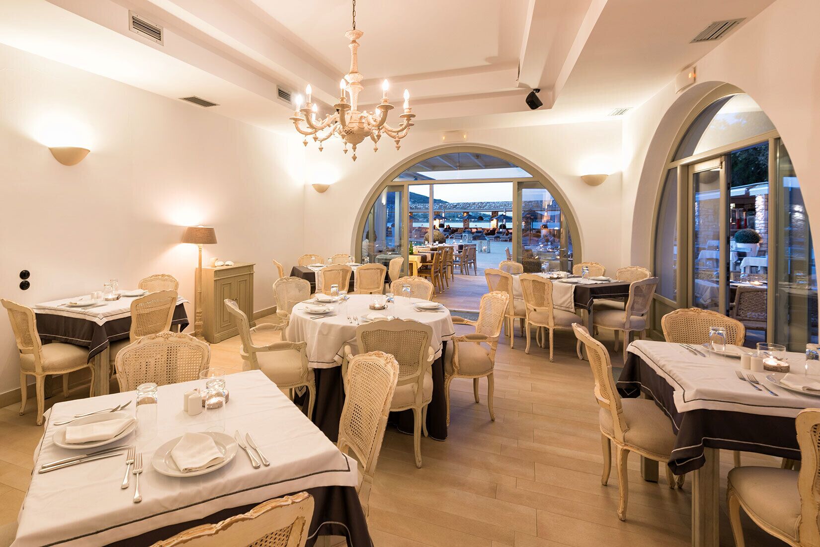 A restaurant with round tables, wicker chairs, and a chandelier.