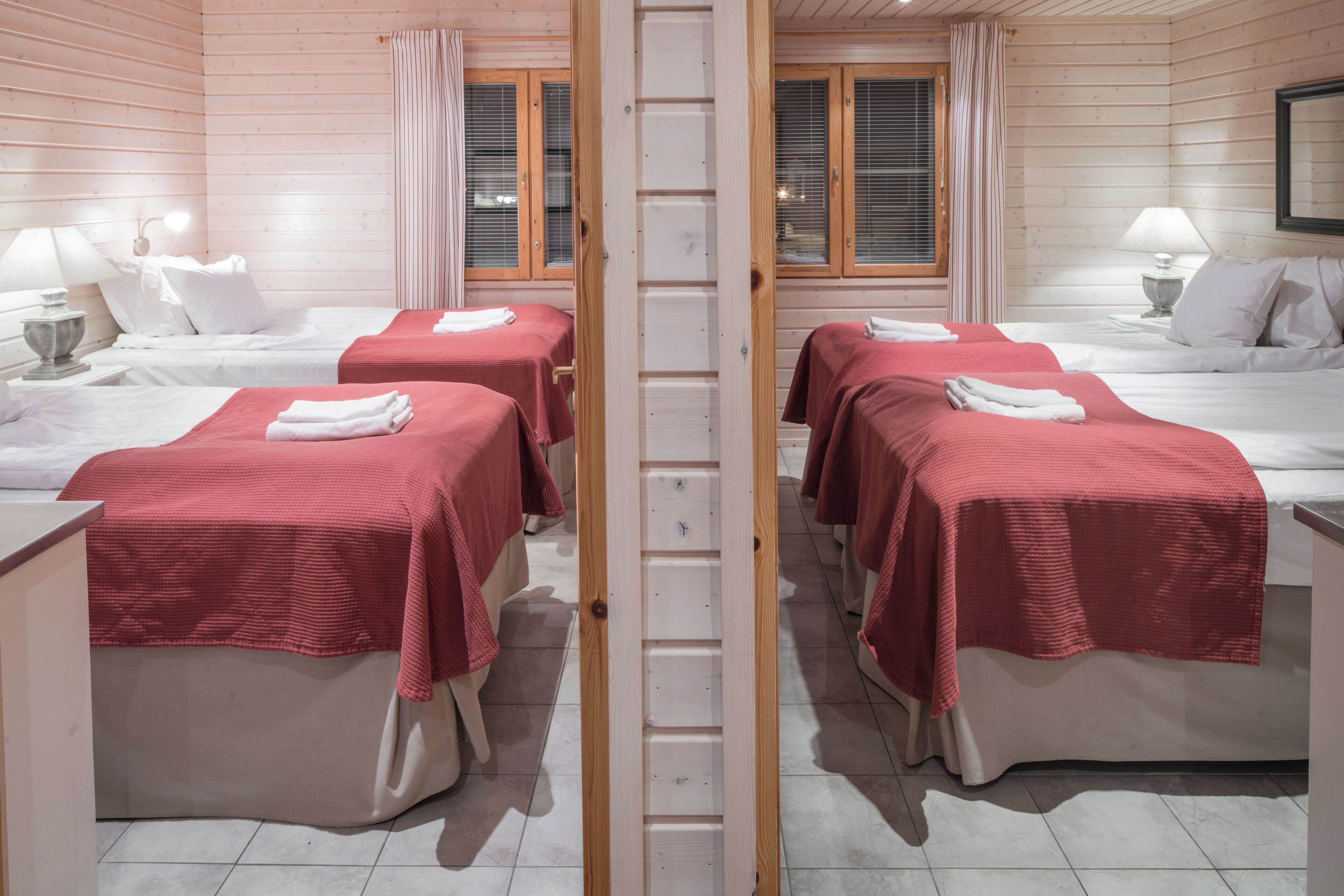 Ein Zimmer mit vier Betten, jedes mit einer roten Bettdecke und weißen Kissen. In der Mitte steht eine Holzleiter, und die Wände sowie die Fenster mit Vorhängen sind aus Holz.
