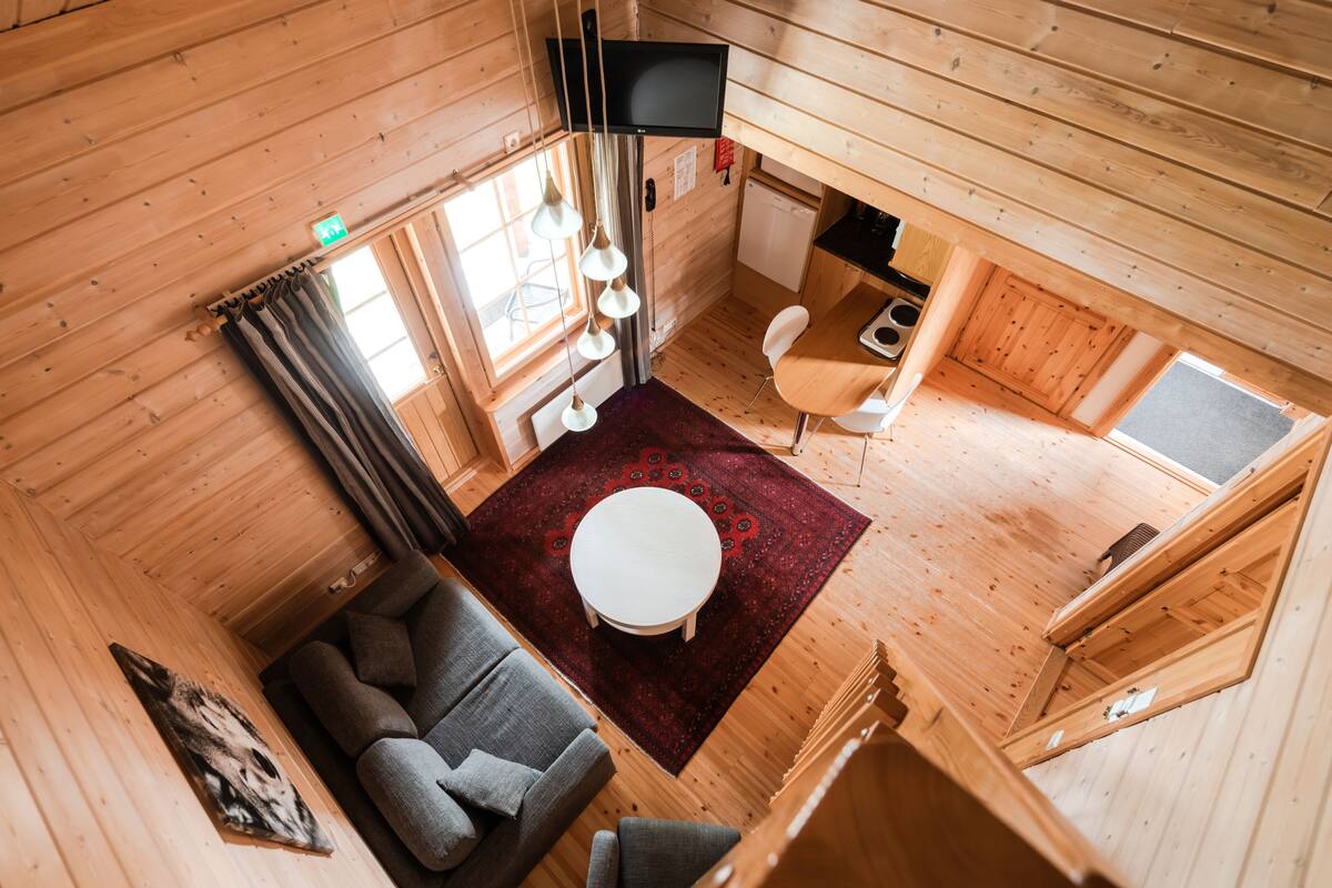 Interior de una cabaña de madera con un sofá, una mesa redonda, una zona de cocina y un televisor montado en la pared.