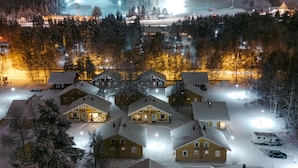 Ein verschneites Dorf mit Holzhäusern, einer Seilbahn und einer beleuchteten Skipiste bei Nacht.