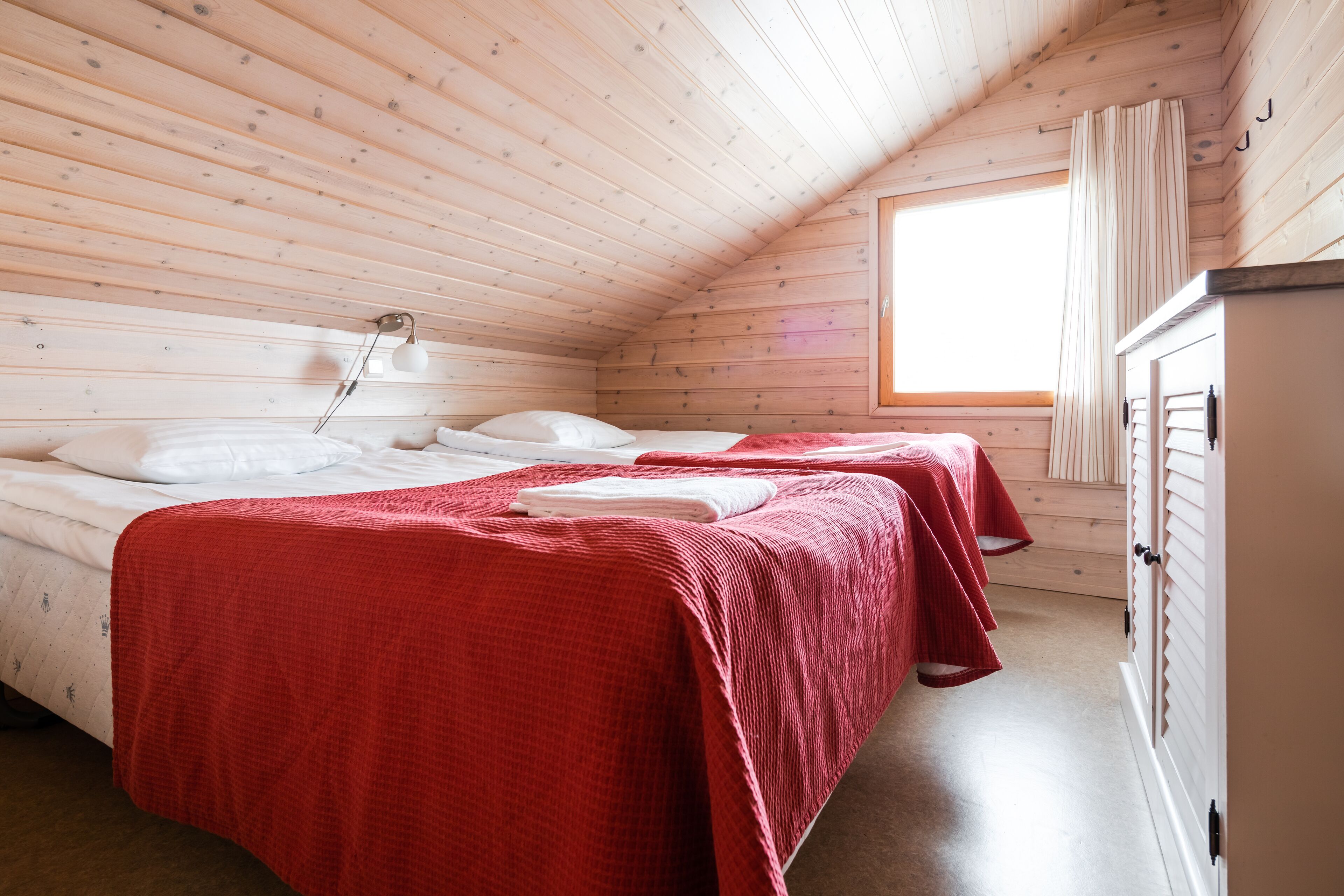 Ein gemütliches Blockhauszimmer mit zwei Betten, Holzboden, einem Fenster mit Vorhängen und einem Kleiderschrank.