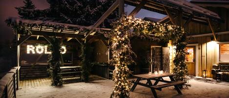Un espacio exterior cubierto de nieve con una estructura de madera, un banco y un árbol de Navidad decorado con luces.