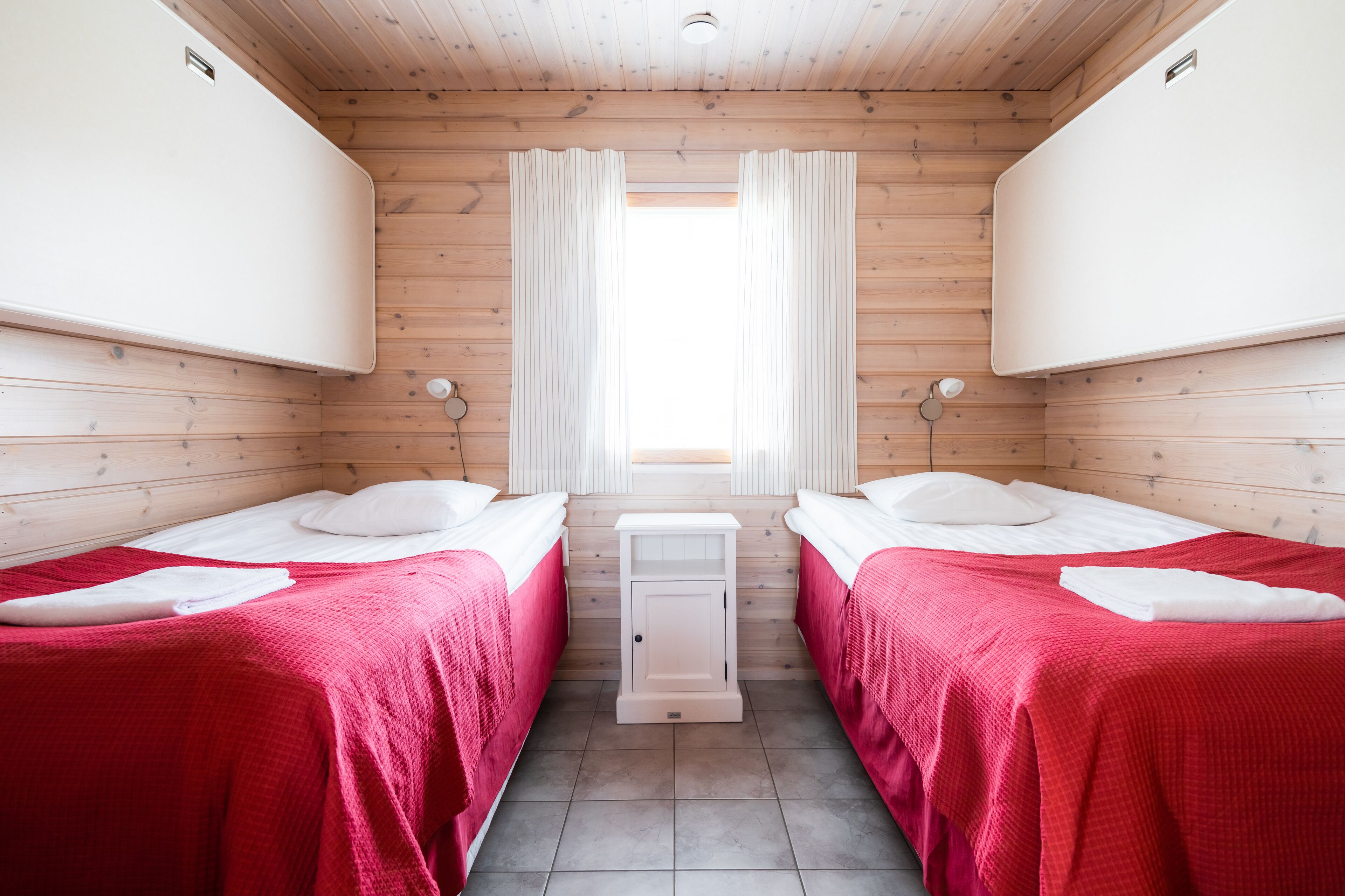 Une chambre avec deux lits, chacun recouvert d’une couverture rouge et agrémenté d’oreillers blancs, une petite table de chevet blanche et une fenêtre avec des rideaux.