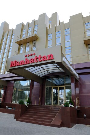 L’entrée d’un hôtel, avec une enseigne « Manhattan » bien visible, de grandes fenêtres en verre et une architecture moderne.