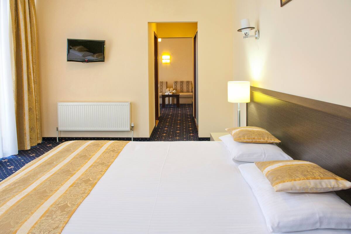 A hotel room with a large bed, a bedside table, a lamp, a view of the hallway, and a radiator.