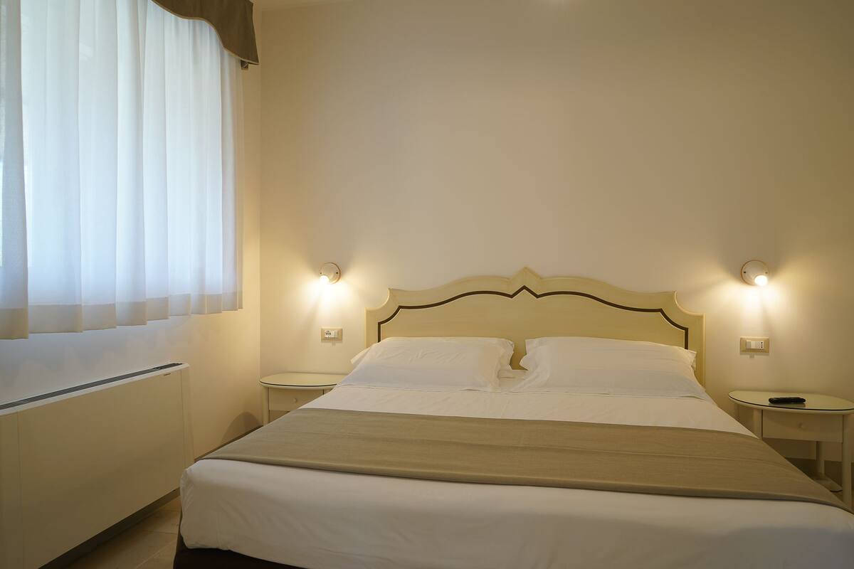 Un letto rifatto con lenzuola bianche e una coperta beige, due comodini con lampade, un termosifone e una finestra con tende trasparenti.
