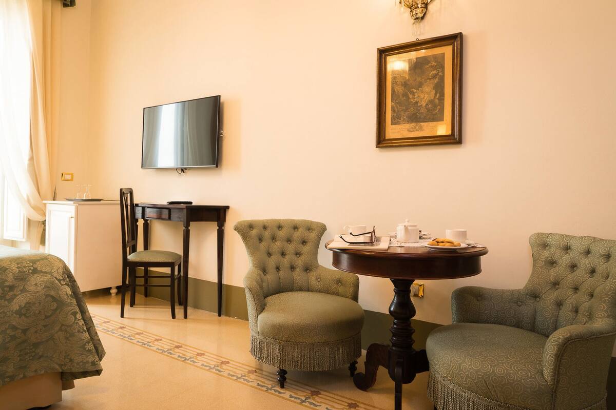 Una camera d'albergo con un letto, due poltrone verdi, un tavolino rotondo con tazze e un teiera, una televisione a parete e un quadro incorniciato sopra il tavolo.