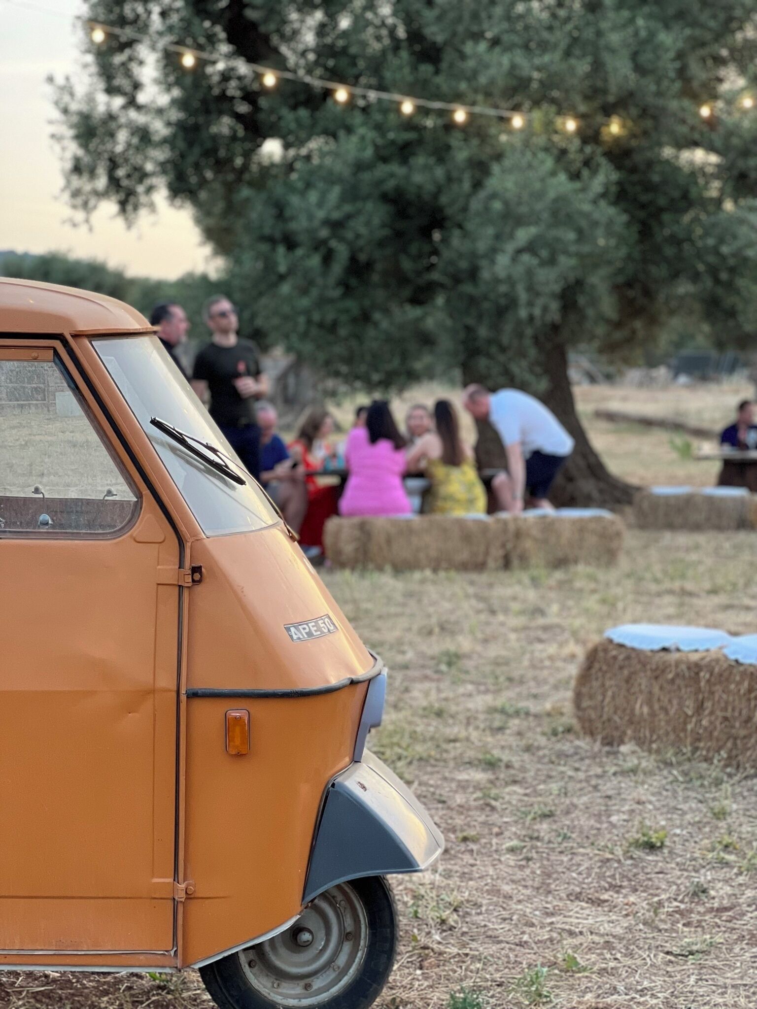 A vintage orange car parked in a rural setting with hay bales and a tree in the background.