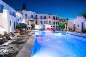Una piscina con sillas de descanso y sombrillas, rodeada de edificios blancos con detalles azules.