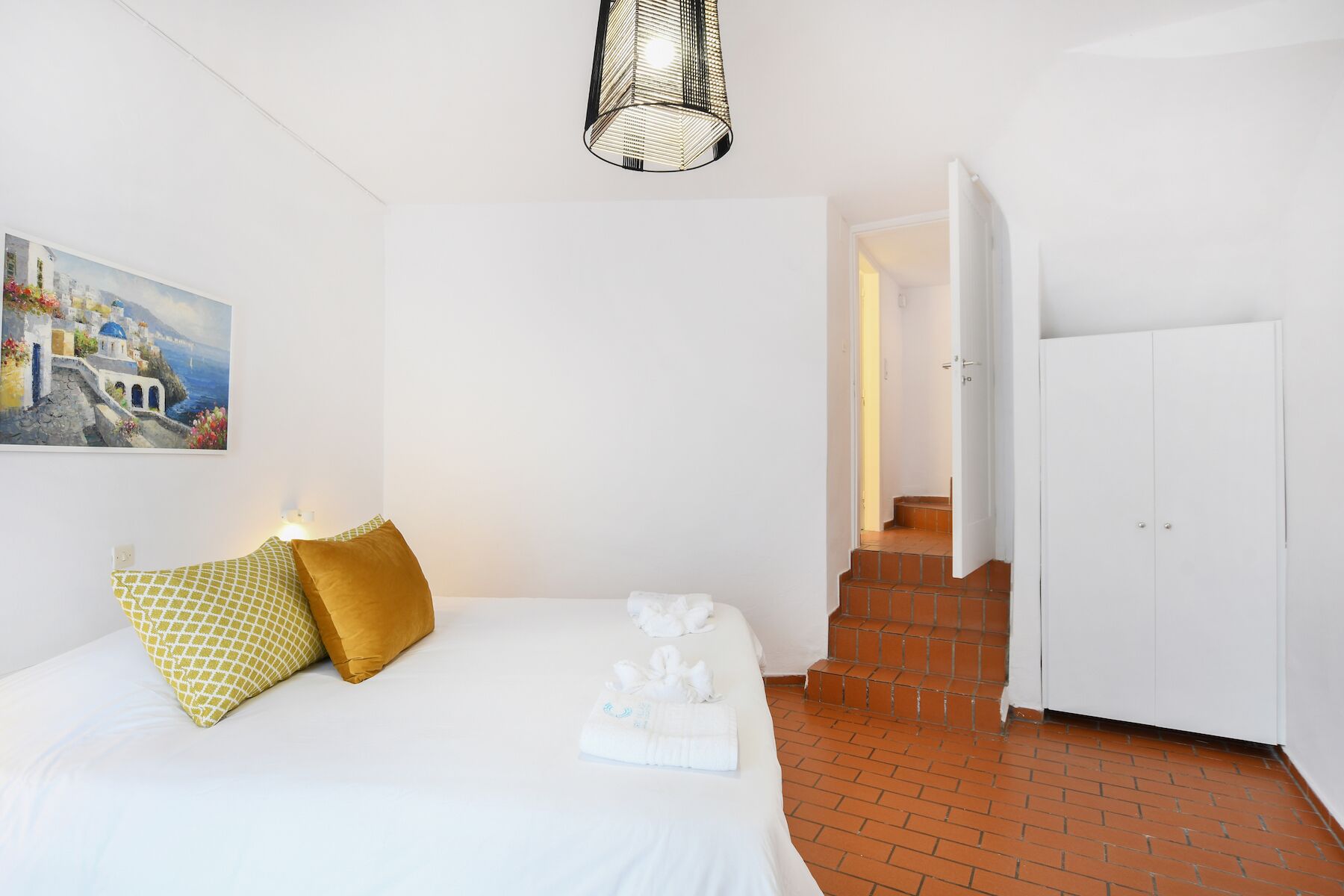 A hotel room with a bed, a painting of a coastal scene, a staircase, and a white door.