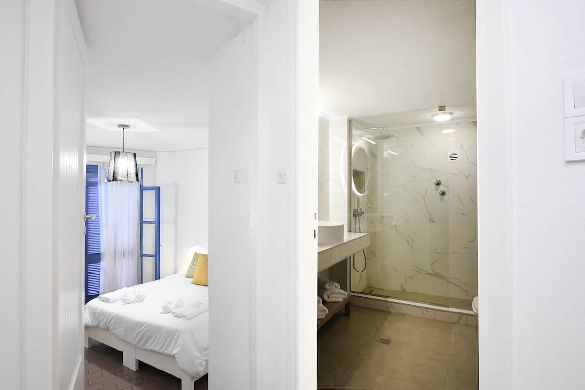 A hotel room with a bed, a bathroom with a glass shower enclosure, and a blue-framed window.