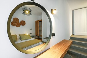 Un dormitorio con un espejo circular, una cama con almohadas estampadas, una mesita de noche y un banco de madera.