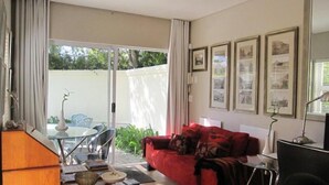 A living room with a red sofa, a glass table, and chairs. There are framed pictures on the wall and a sliding glass door leading to an outdoor area. - Navona Constantia Guest Cottage (Cape Town)