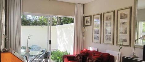 A living room with a red sofa, a glass table, and chairs. There are framed pictures on the wall and a sliding glass door leading to an outdoor area.