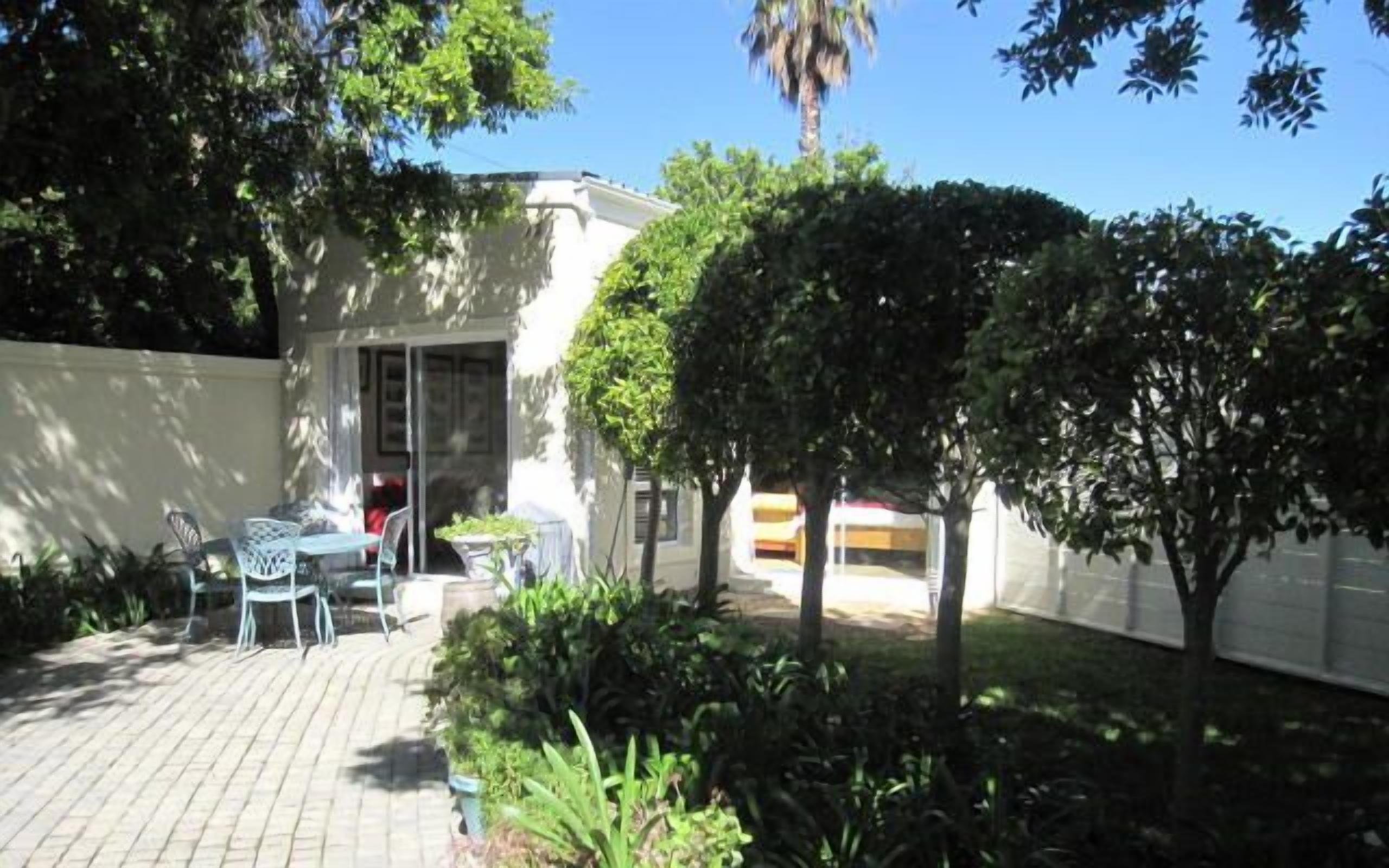 A patio area with a table and chairs, surrounded by greenery and a white building.