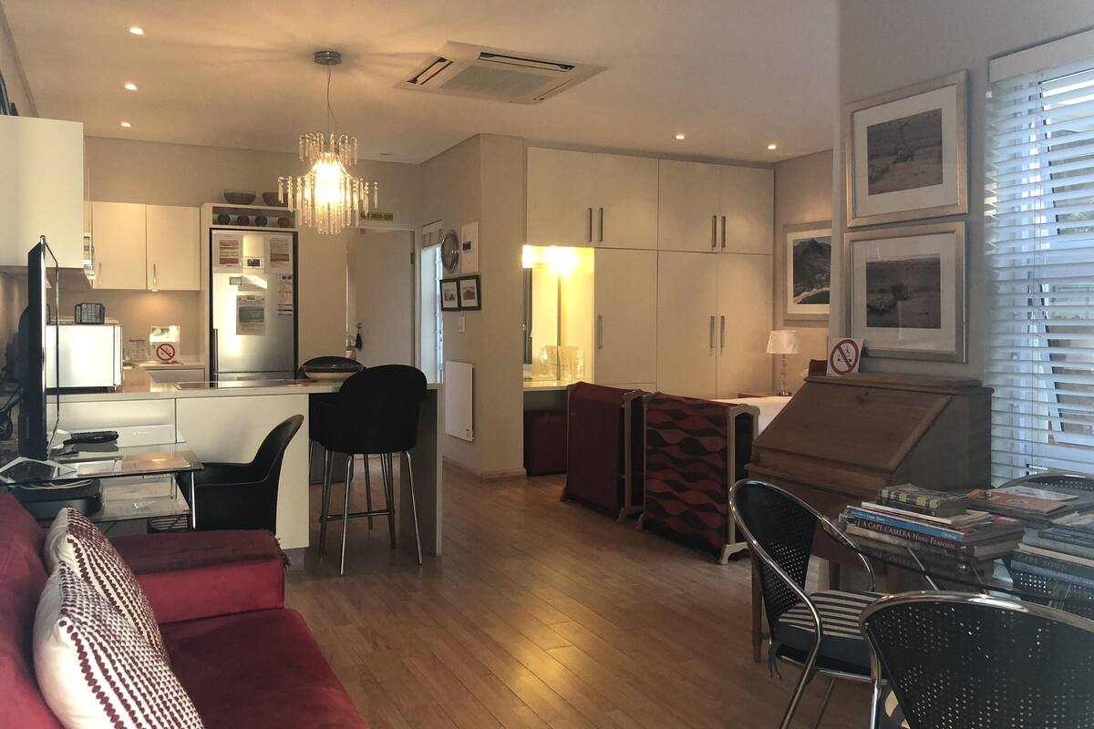 A modern living room with a red sofa, a dining area with a table and chairs, a kitchen with white cabinets, and a chandelier.
