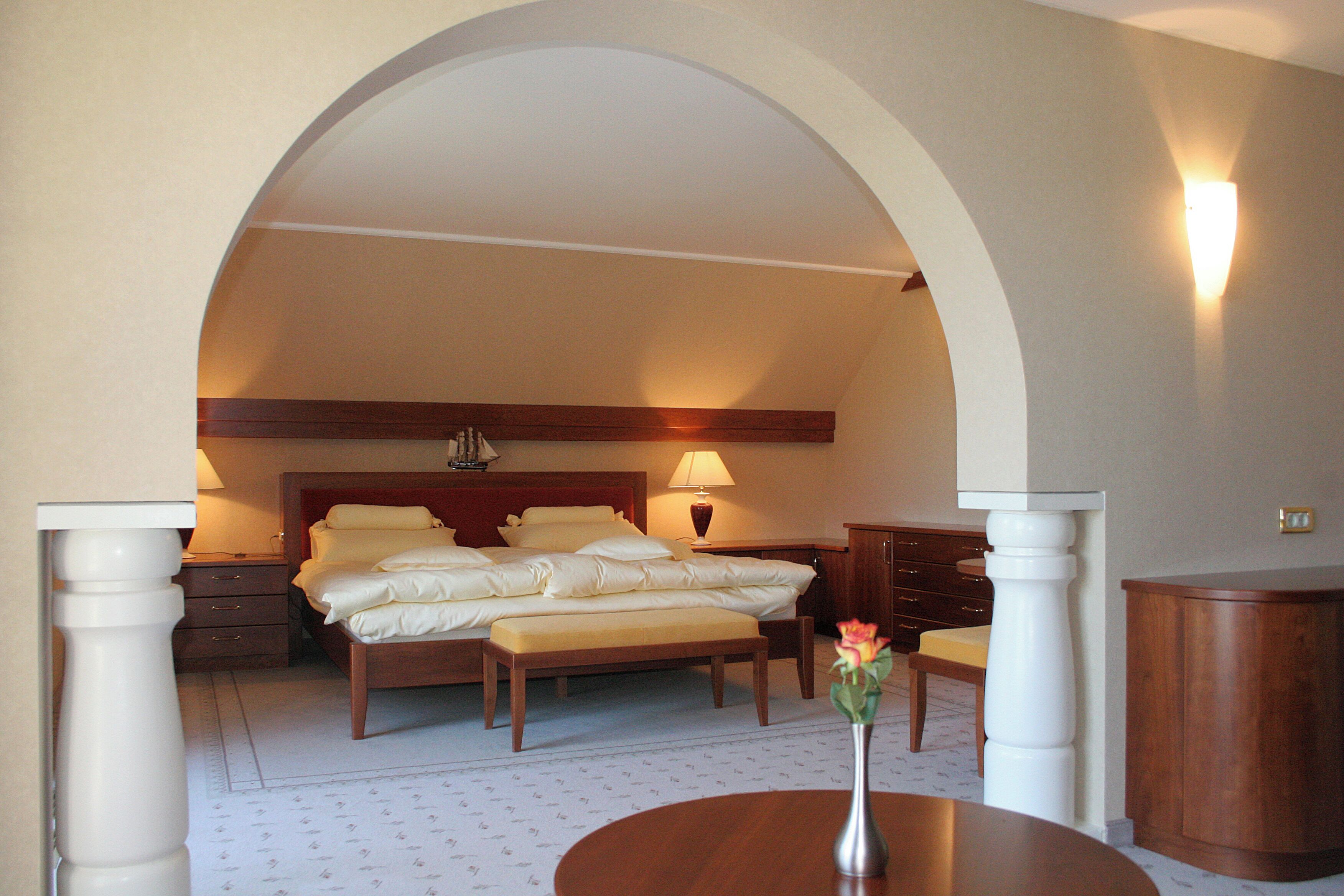 Habitación de hotel con un amplio arco de entrada, cama con sábanas blancas, mesita de noche con lámpara, banco de madera y un jarrón con flores.