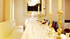Ein langer Tisch ist mit weißen Tischdecken, goldenen Kerzenständern und weißen Stühlen in einem Raum mit cremefarbenen Wänden und einem Kronleuchter gedeckt.